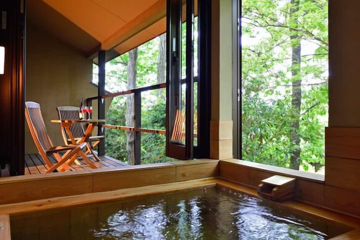 A wooden interior with a hot tub, wooden chairs, and a view of trees through large windows.