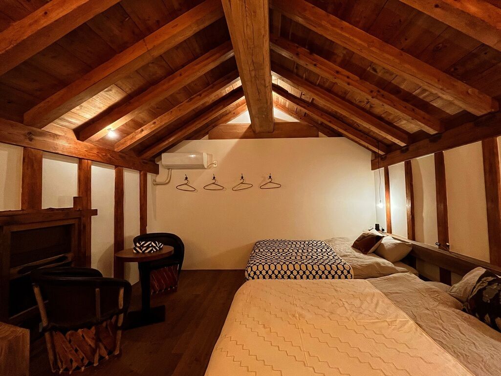 An attic bedroom with exposed wooden beams, a bed with patterned bedding, a chair, and a small table.