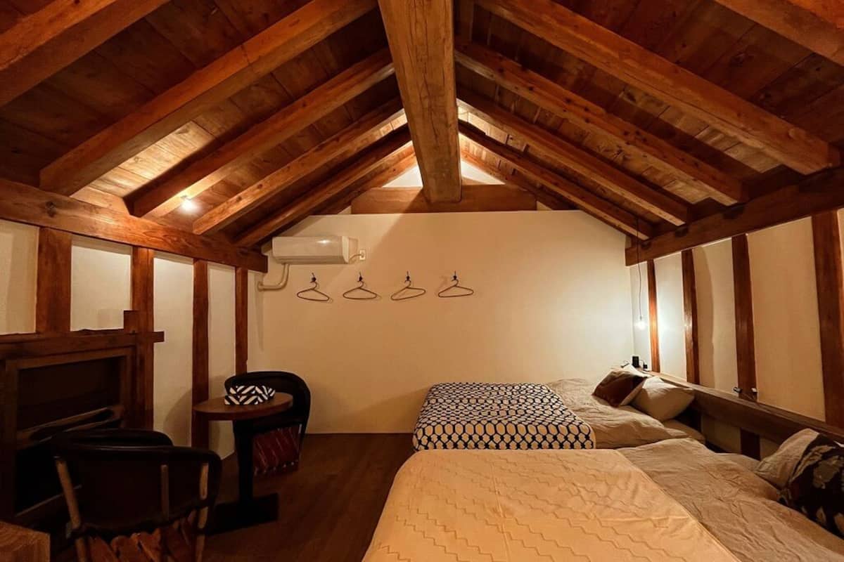 An attic bedroom with exposed wooden beams, a bed with patterned bedding, a chair, and a small table.