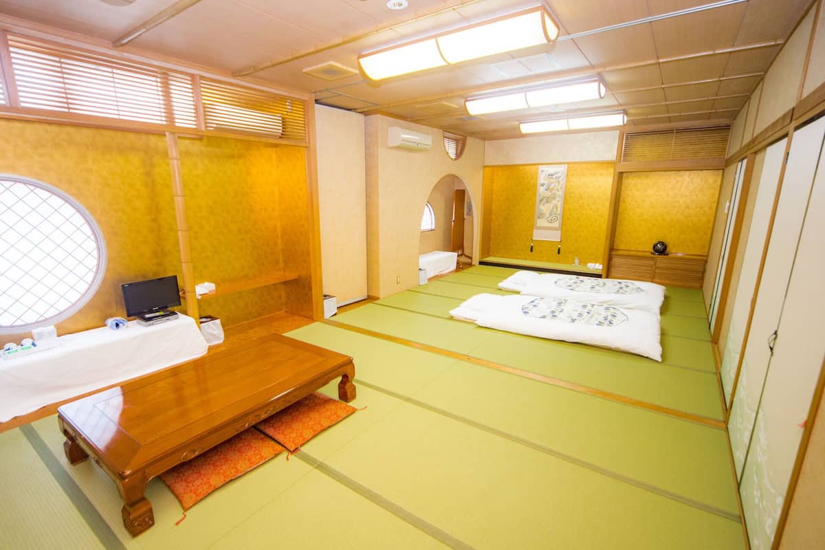 A traditional Japanese room with tatami flooring, a futon bed, a wooden desk with a computer, and a window with blinds.