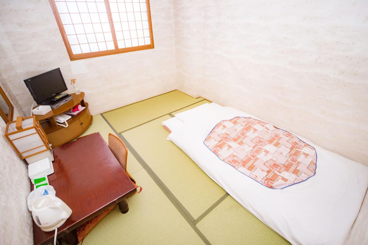 A room with tatami flooring, a bed with a patterned blanket, a desk with a computer, and a small table with a chair.