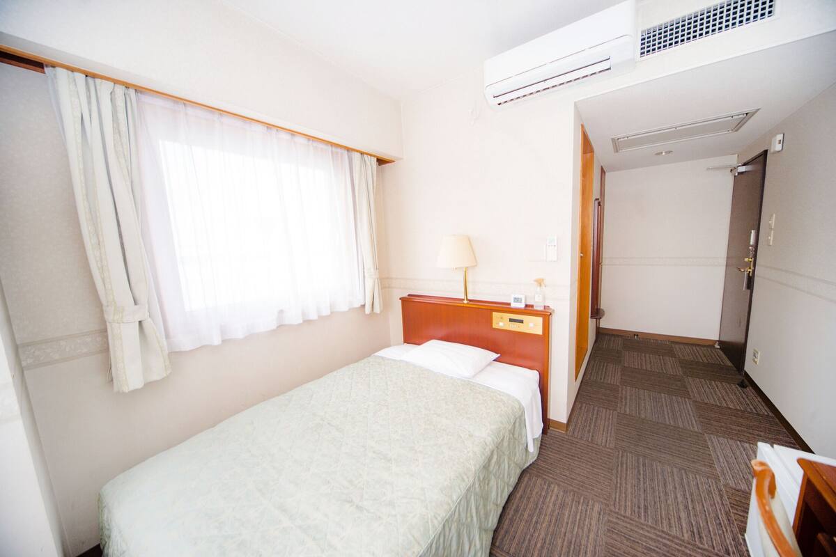 A hotel room with a bed, a nightstand, a lamp, an air conditioning unit, and a door leading to another room.