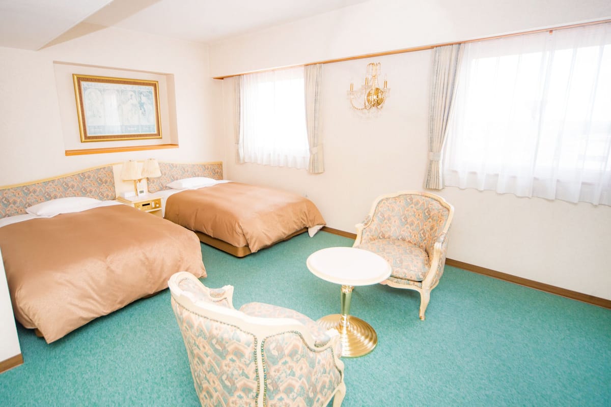 A hotel room with two beds, a small round table, a chair, a chandelier, and a framed picture on the wall.