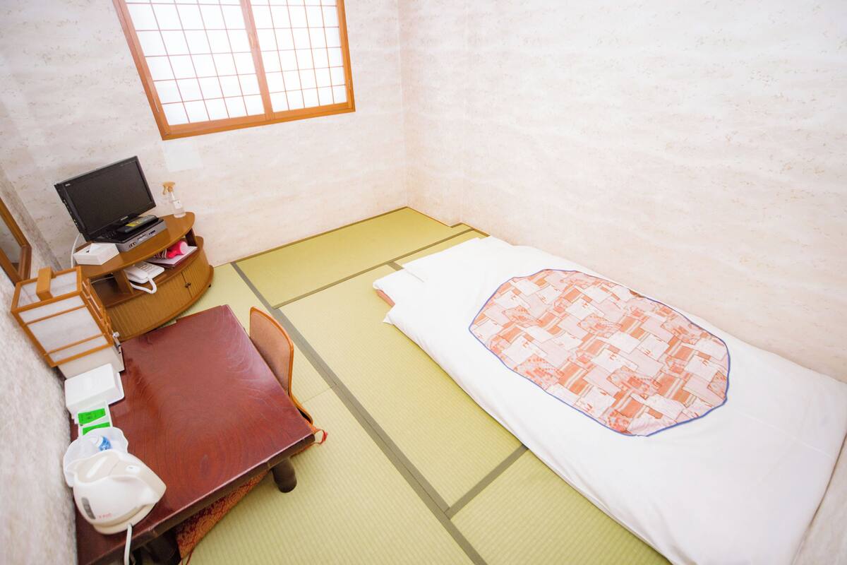 A room with tatami flooring, a bed with a patterned blanket, a desk with a computer, and a small table with a chair.