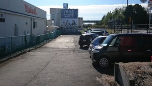 Un estacionamiento con varios coches, un edificio con un cartel que dice 'Yakuji' y un gran cartel publicitario al fondo.