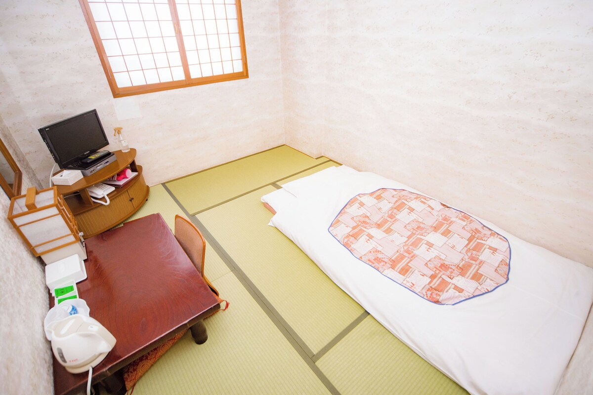Una habitación con suelo de tatami, una cama con manta estampada, un escritorio con computadora y una pequeña mesa con silla.