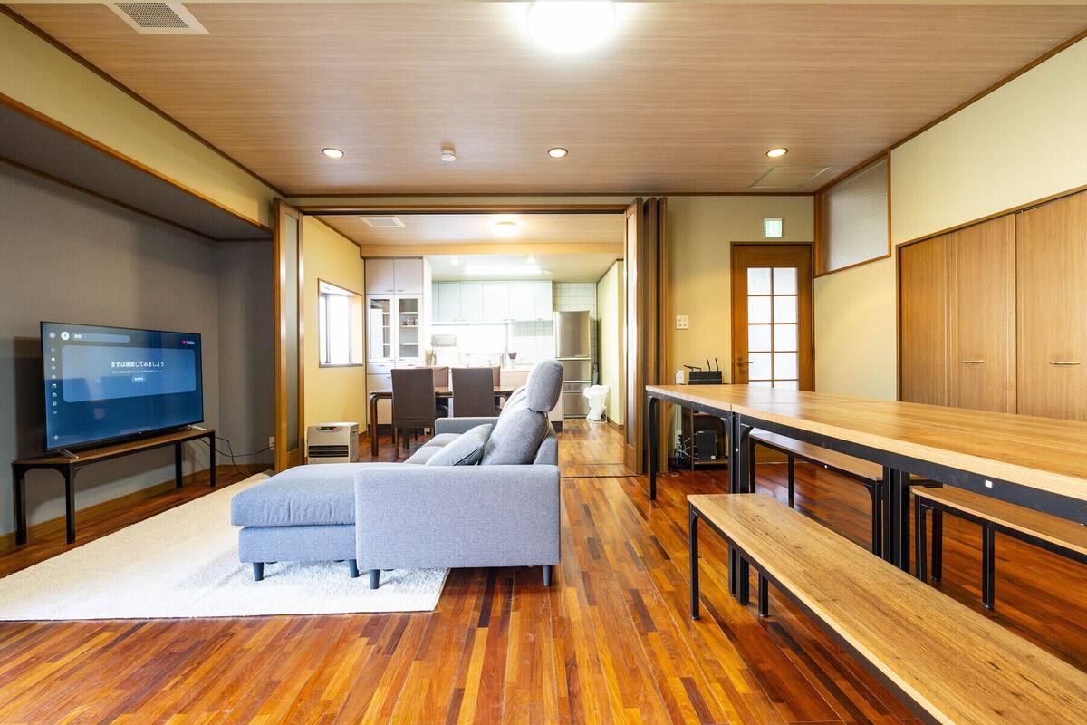 A modern living room with a flat-screen TV, grey sofa, wooden benches, and a kitchen in the background.