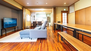 A modern living room with a flat-screen TV, a grey sofa, wooden flooring, and a dining area with benches. - Oyado Sakura Gojo Station (Gojo)