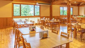 A spacious dining area with multiple wooden tables and chairs, large windows with a view of greenery, and an air conditioning unit on the ceiling.