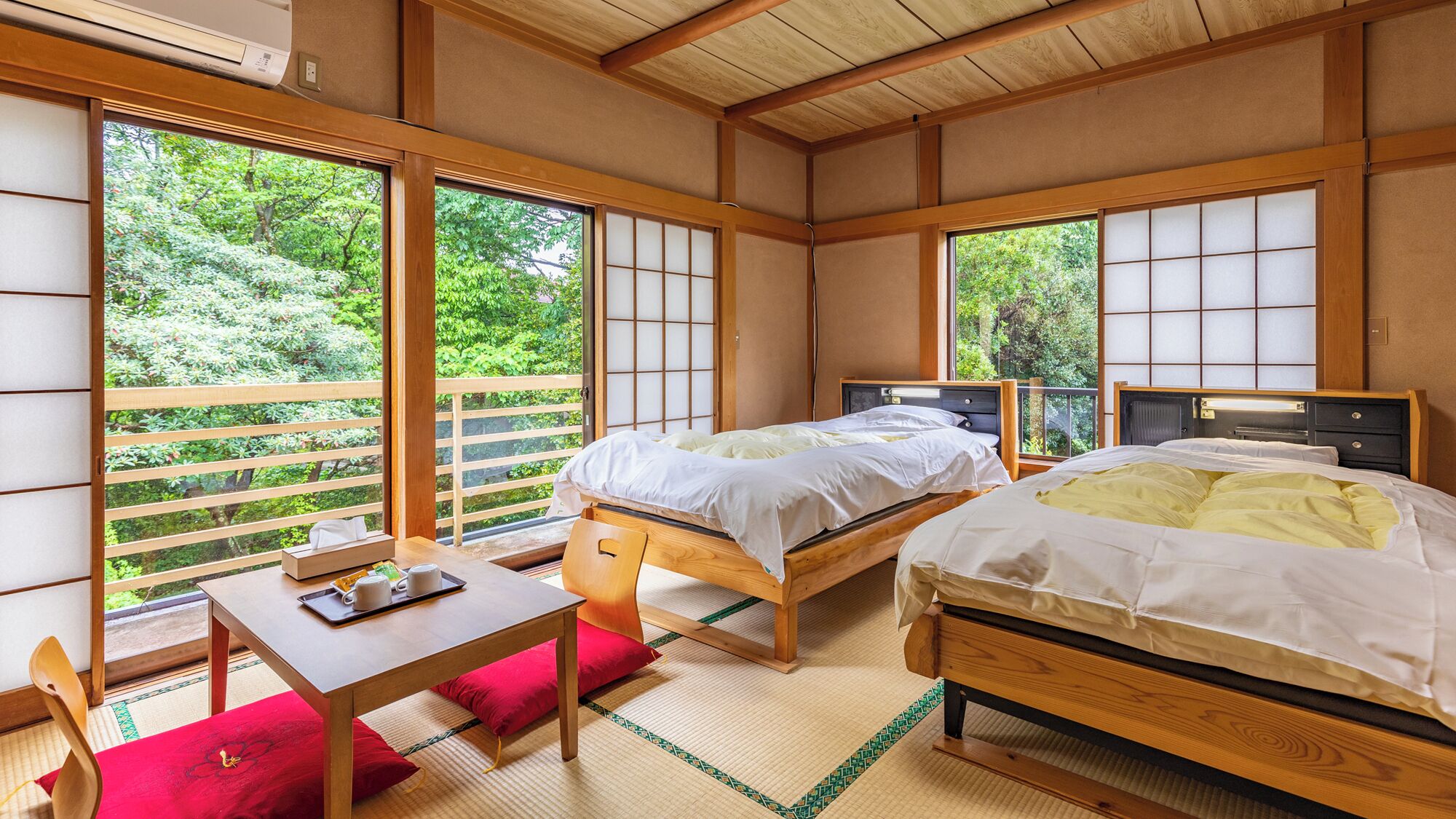 Una habitación tradicional japonesa con suelo de tatami, dos camas de madera, una mesita con una bandeja de objetos y ventanas amplias que ofrecen vistas a la vegetación.