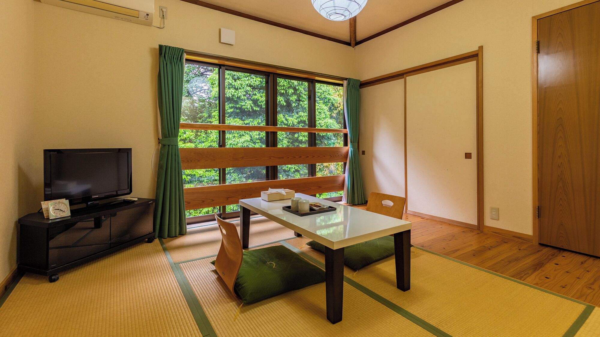 Una habitación con suelo de tatami, un televisor sobre un mueble, una mesa de madera con cojín y una puerta corredera.