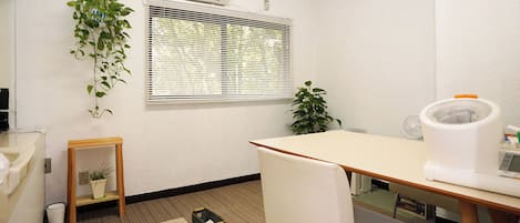 Ein kleines Büro mit Schreibtisch, Stuhl, Fenster mit Jalousien, Klimaanlage und Topfpflanzen.