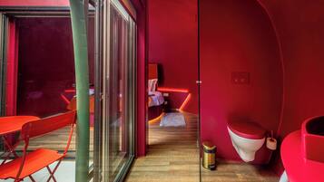 Habitación moderna con puerta de vidrio que da a un espacio al aire libre, cama con pared de acento roja, una alfombra pequeña y un inodoro con asiento rojo.