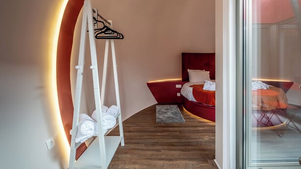 Ett modernt hotellrum med en böjd vägg, en säng med röda sängkläder, en vit hängare för handdukar och en glasdörr som leder ut till en balkong.