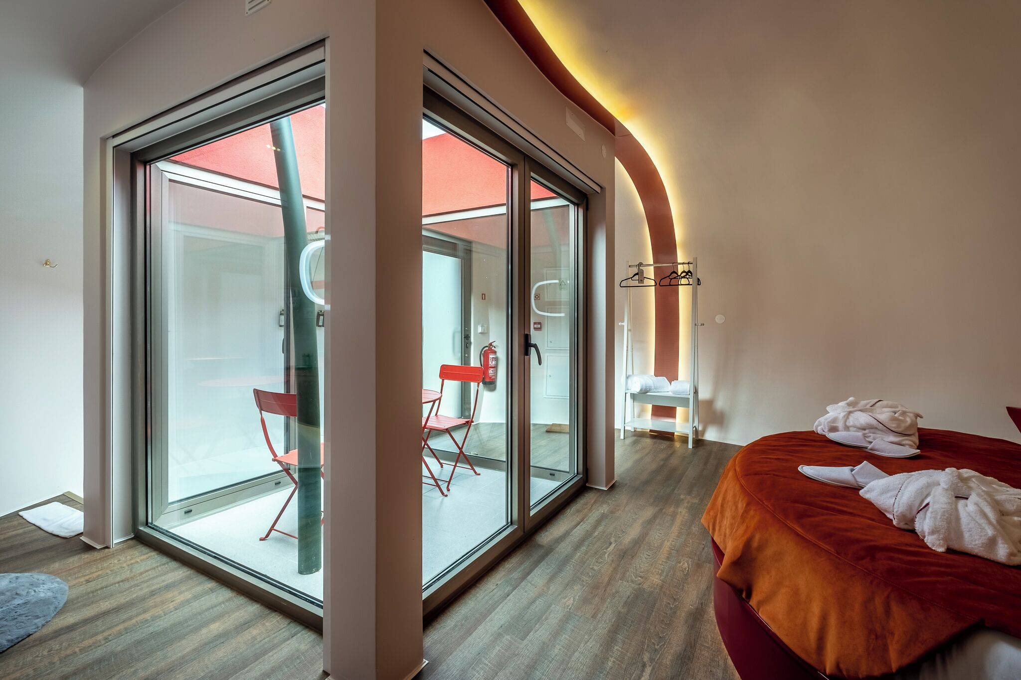 A modern hotel room with a large sliding glass door leading to a balcony, a bed with a red headboard, and a small seating area with a red chair and a white table.