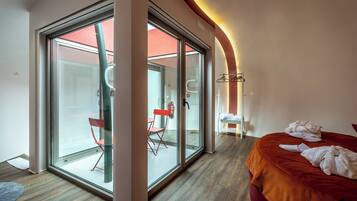 A modern hotel room with a large sliding glass door leading to a balcony, a bed with a red headboard, and a small seating area with a red chair and a white table.