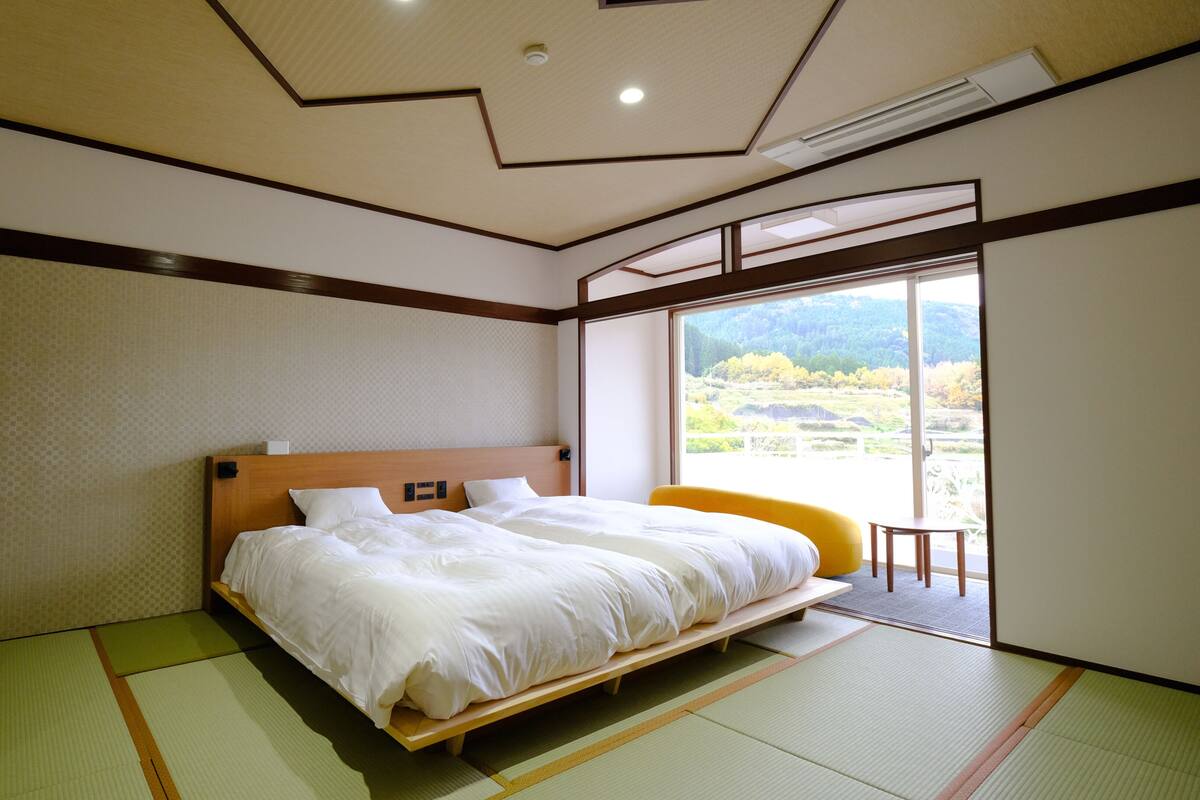 Une chambre d’hôtel avec un grand lit, une vue sur un paysage montagneux, des tatamis au sol et une tête de lit en bois.
