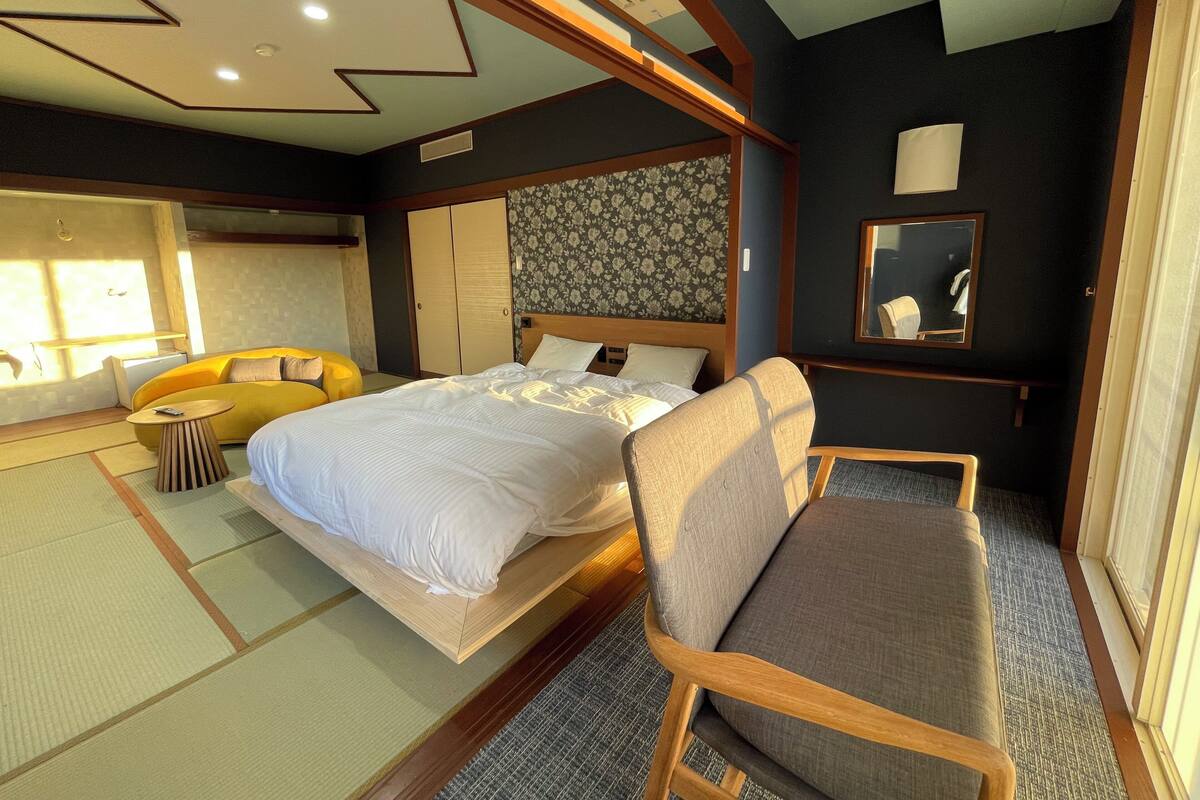 Une chambre d’hôtel avec un lit, un canapé, une chaise, une petite table, un miroir et une fenêtre laissant entrer la lumière du soleil.