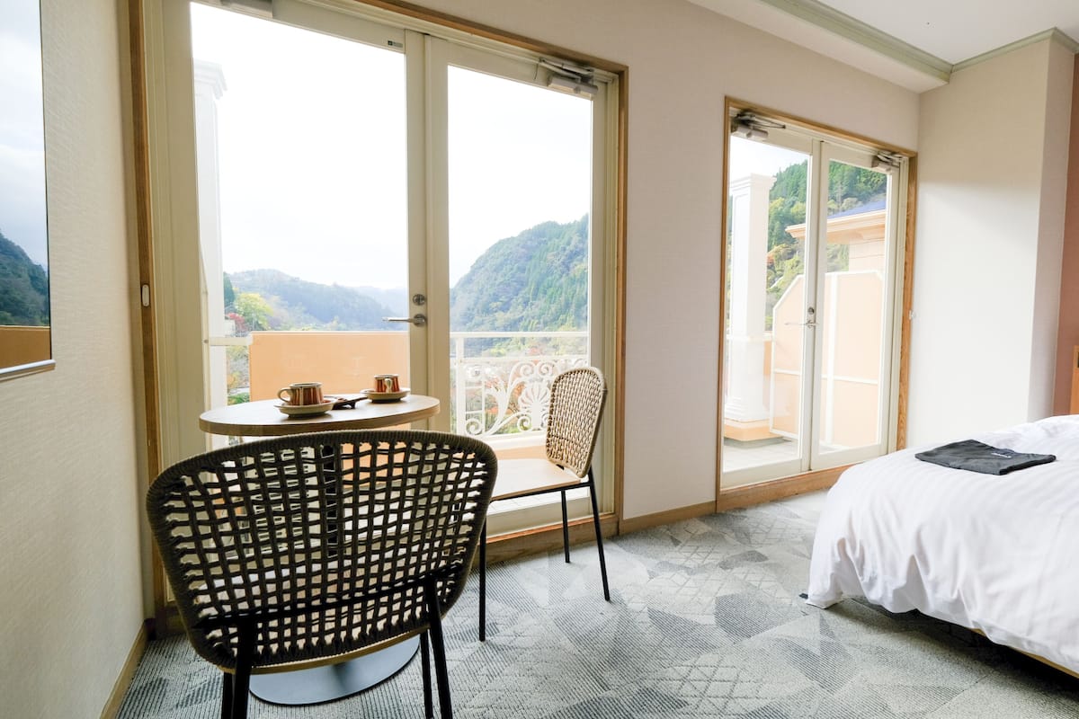 Une chambre d’hôtel avec un lit, une chaise, une table avec deux tasses et un balcon offrant une vue sur les montagnes.