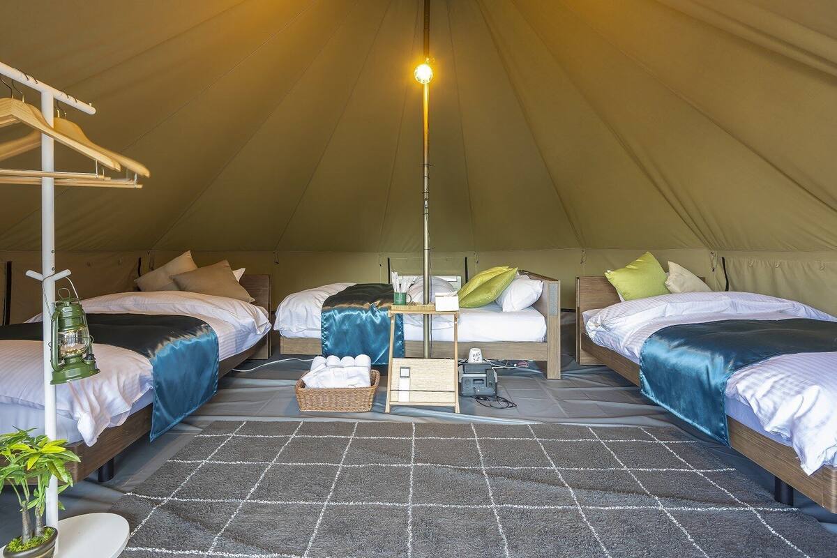 A tented accommodation with two beds, a hanging light, and a small table with a lamp.