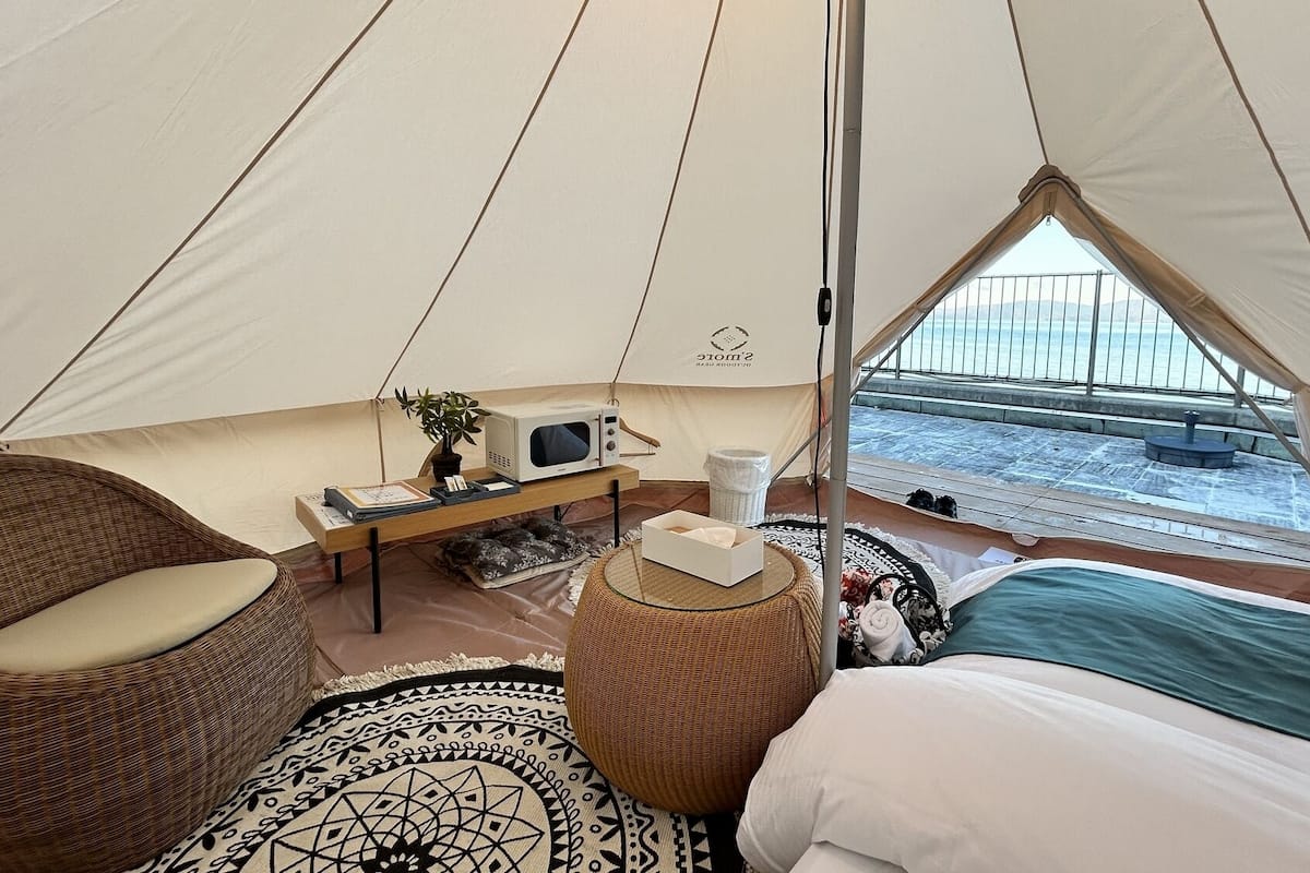 A tent with a bed, a chair, a small table with a plant, a microwave, and a view of a snowy landscape.
