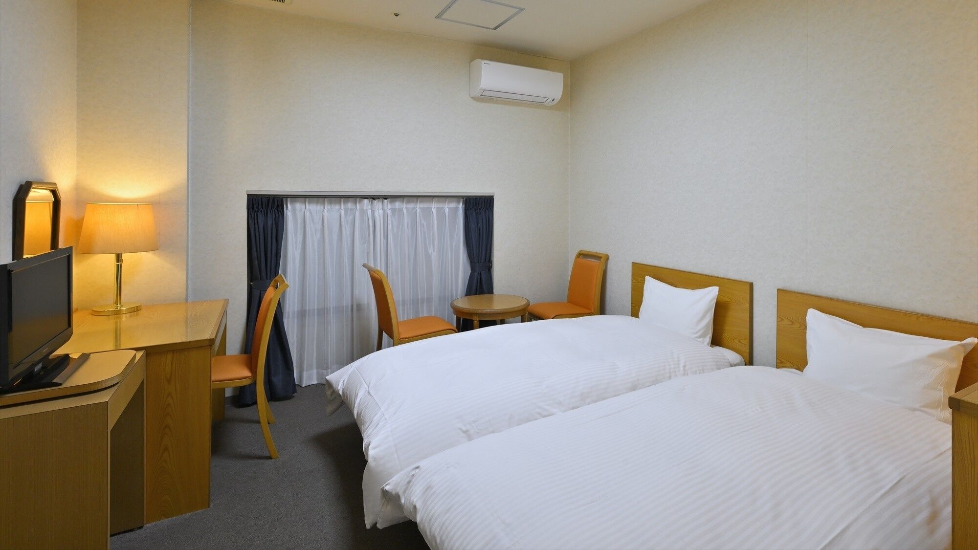 A hotel room with two beds, a desk with a TV, a lamp, a chair, a small table, and an air conditioning unit.