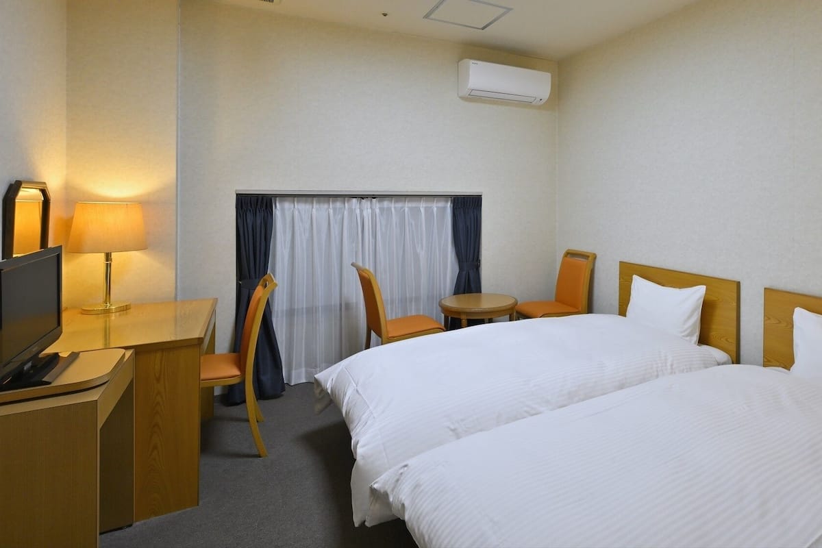 A hotel room with two beds, a desk with a TV, a lamp, a chair, a small table, and an air conditioning unit.