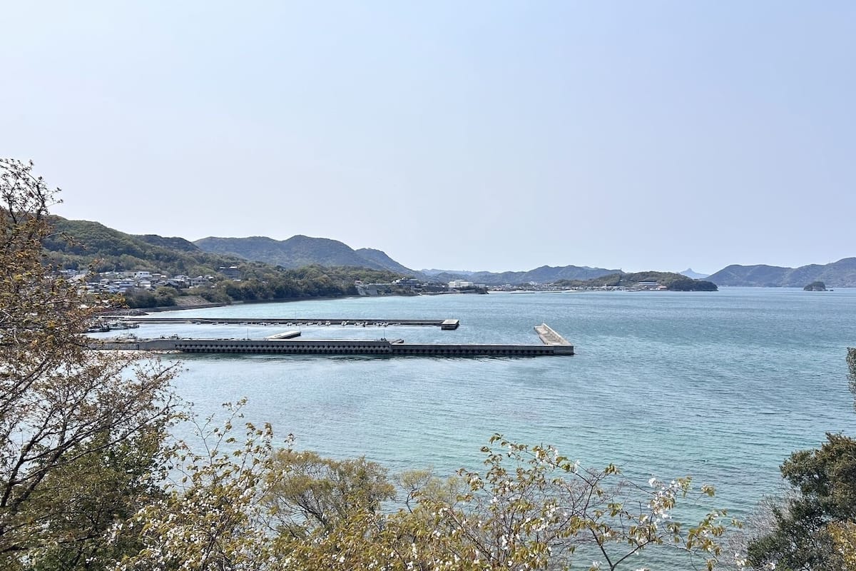 A coastal landscape with a pier extending into calm waters, surrounded by hills and sparse vegetation.