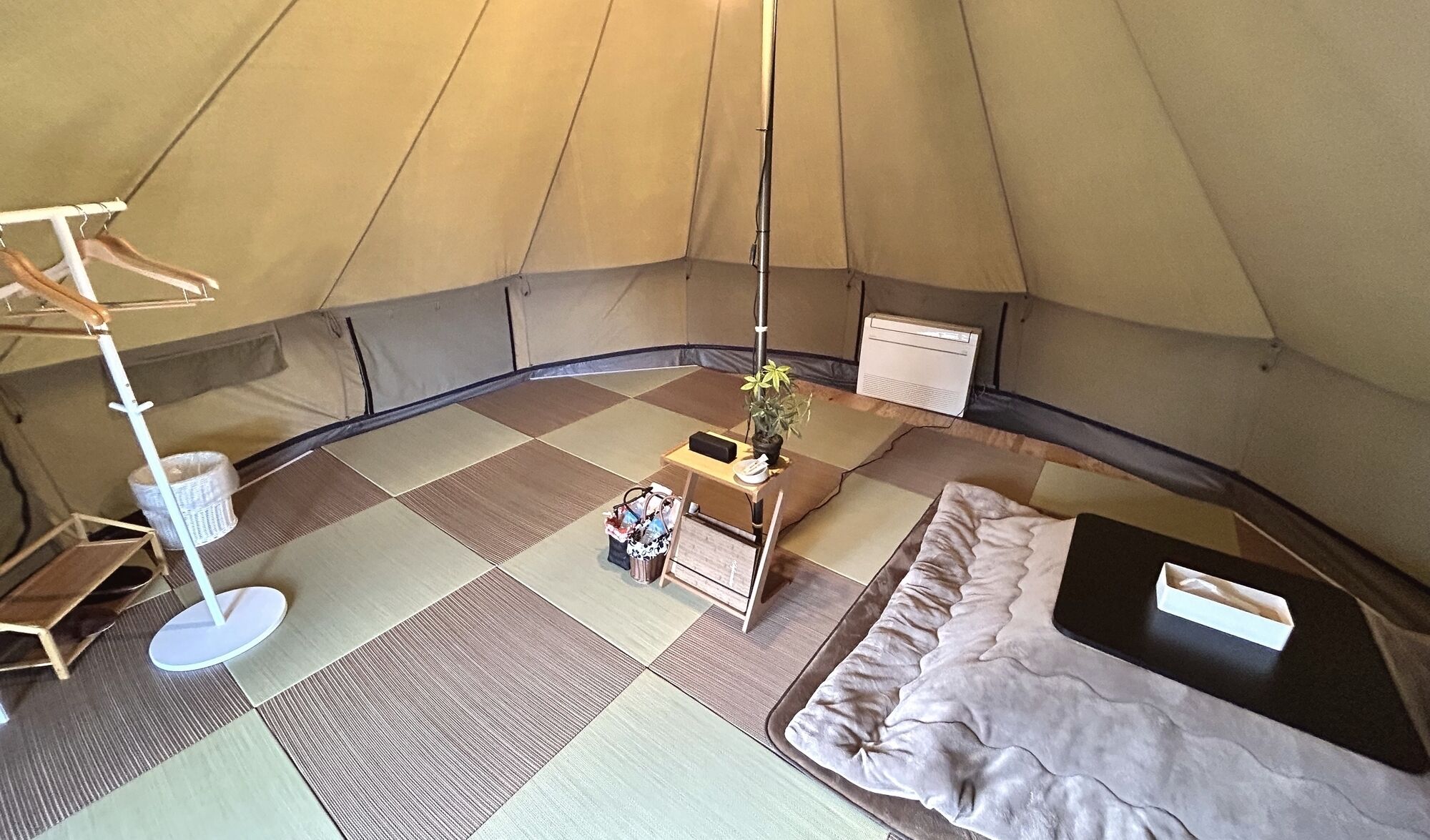 A tent interior with a bed, a table with a plant, a hanging light, and a portable heater.