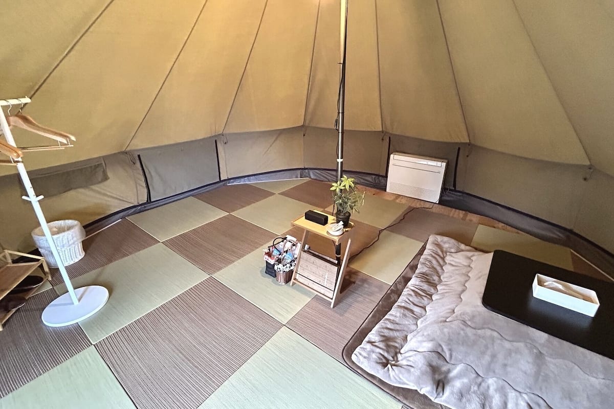 A tent interior with a bed, a table with a plant, a hanging light, and a portable heater.