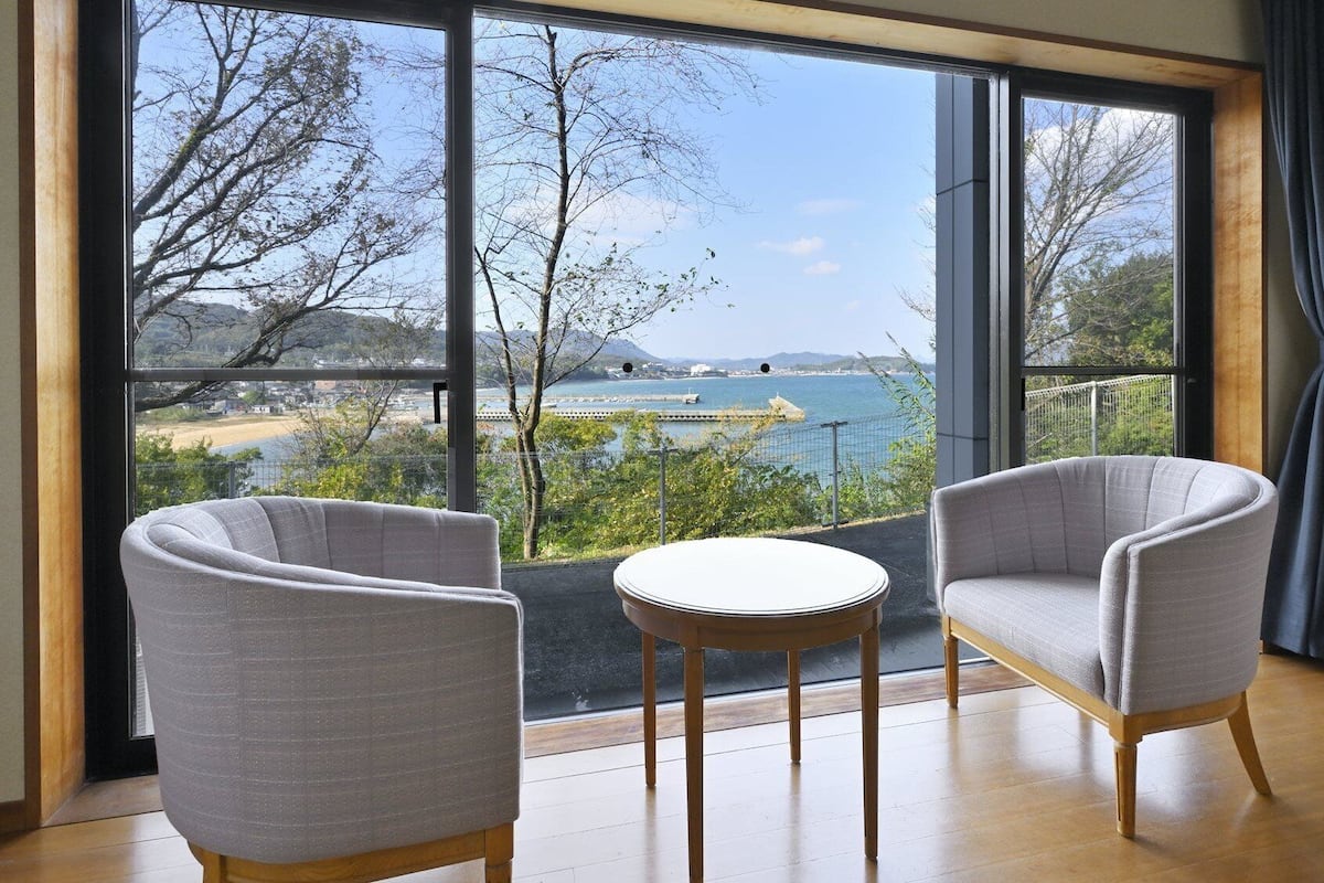 A room with two grey armchairs and a small round table, large windows with a view of a beach and water.