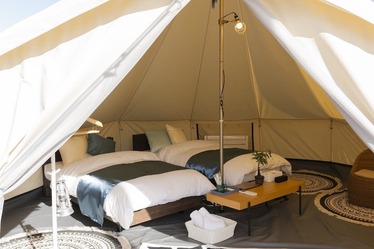 A tent with two beds, a bedside table, a lamp, a chair, and a patterned rug.