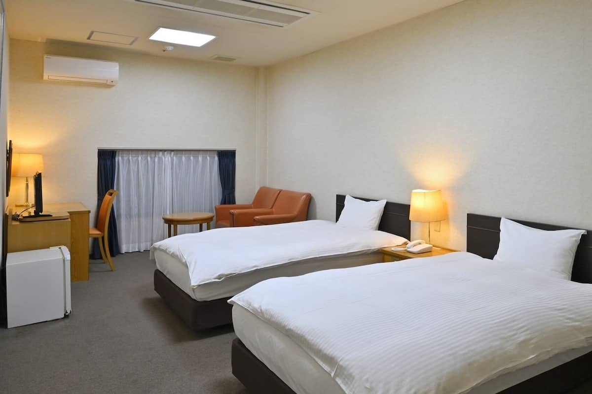 A hotel room with two beds, a desk, a chair, a lamp, and an air conditioning unit.