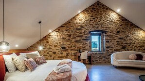 Un dormitorio con pared de piedra, cama con colcha floral, reposapiés rojo, sofá blanco y escritorio con silla.