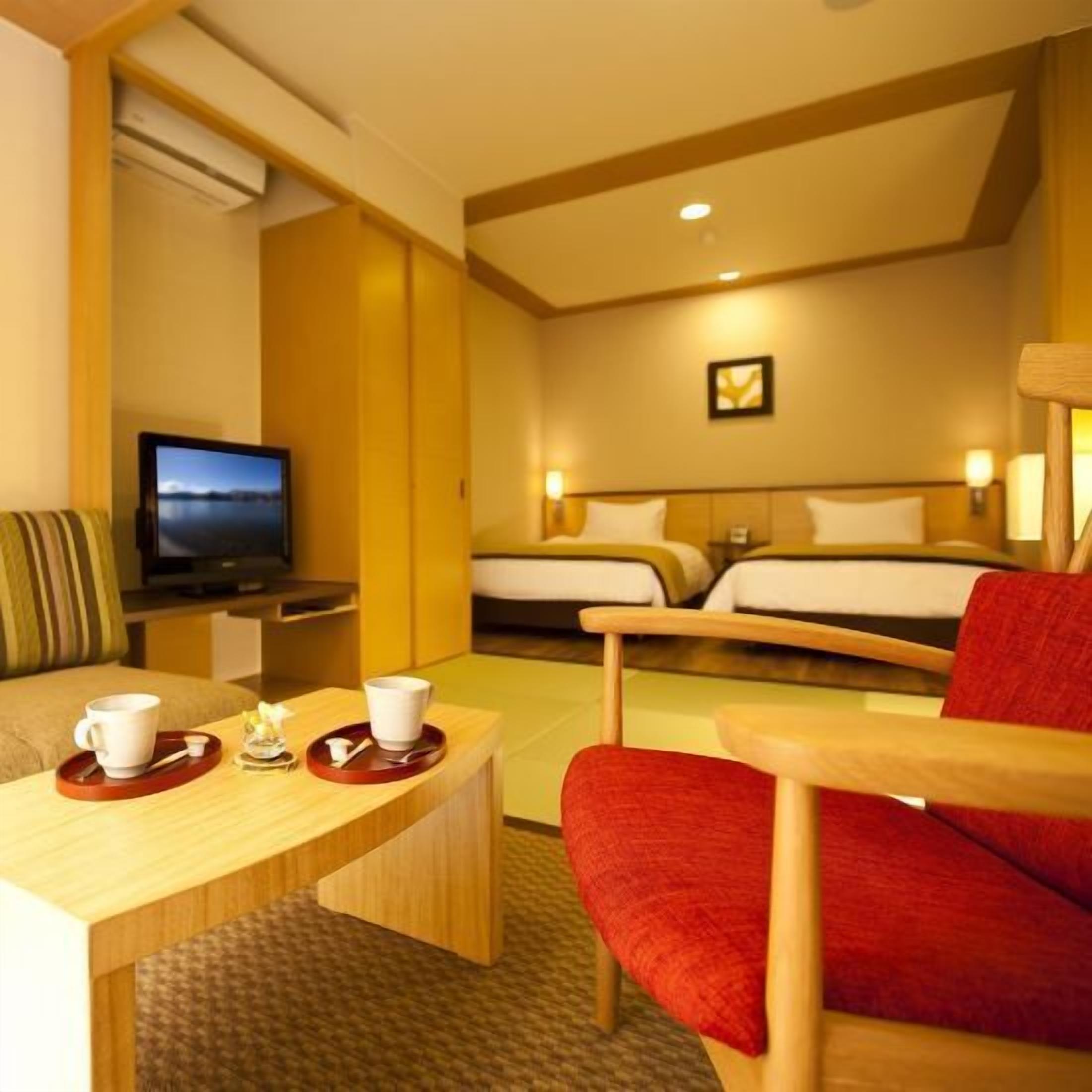 A hotel room with a bed, a TV, a desk with a chair, and a red armchair.