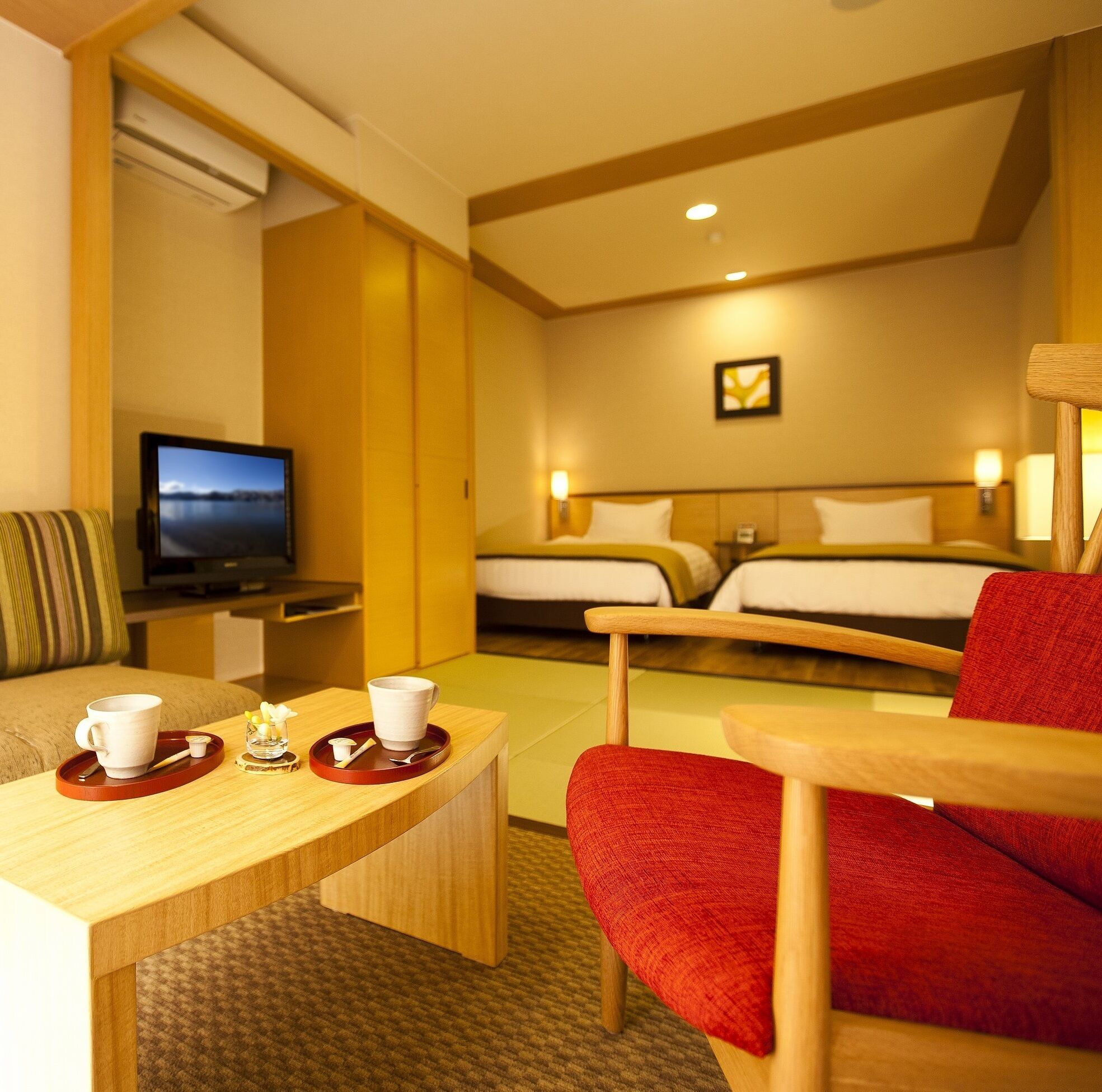 A hotel room with a TV, a desk with a tray, two beds, and a red chair.