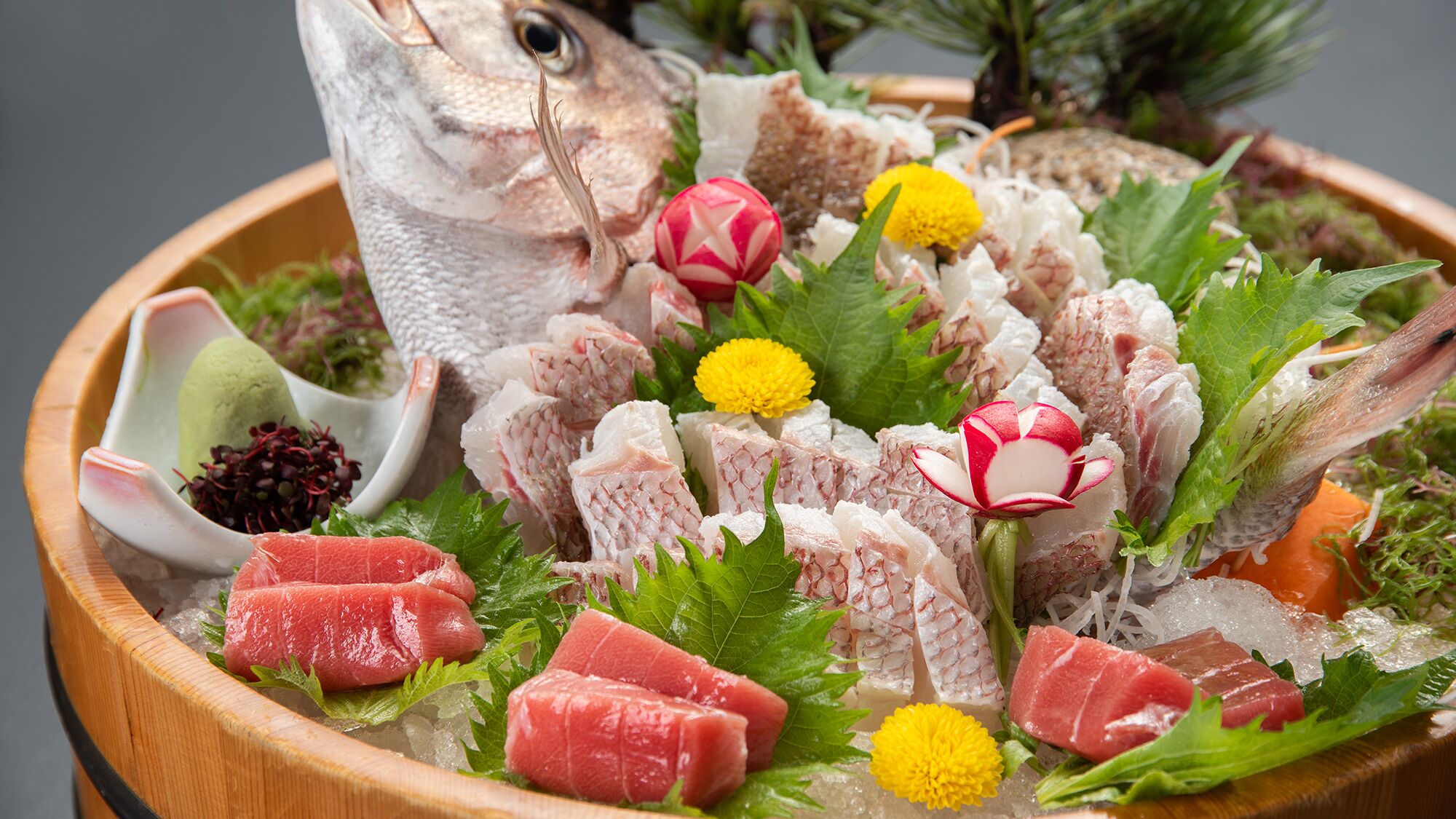 A wooden bowl filled with various types of fish and seafood, garnished with green leaves and flowers.