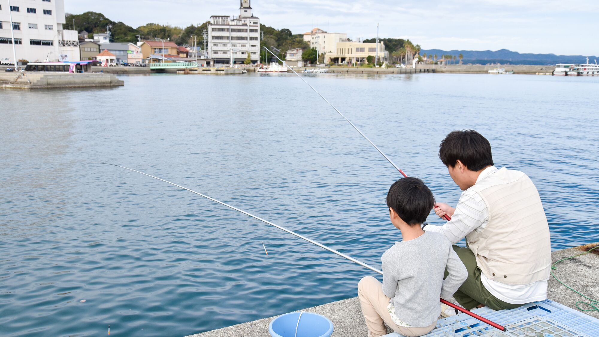 Two people fishing by a calm river with buildings in the background.