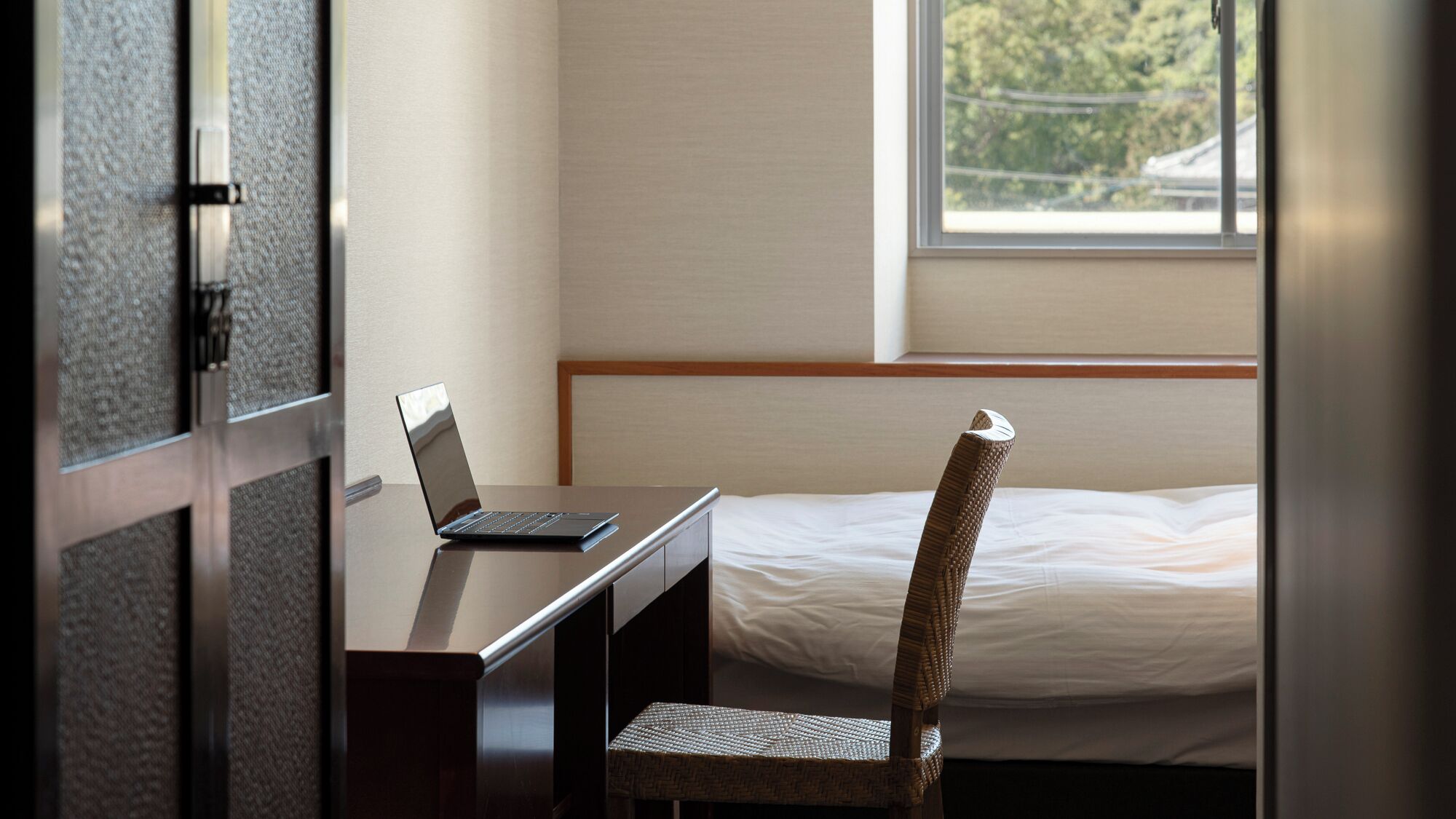Une chambre d’hôtel avec un lit, un bureau avec un ordinateur portable, un fauteuil en osier et une fenêtre donnant sur un paysage verdoyant.