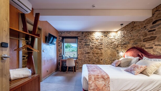 Um quarto com parede de pedra, cama com cabeceira vermelha, guarda-roupa de madeira, escrivaninha e televisão.