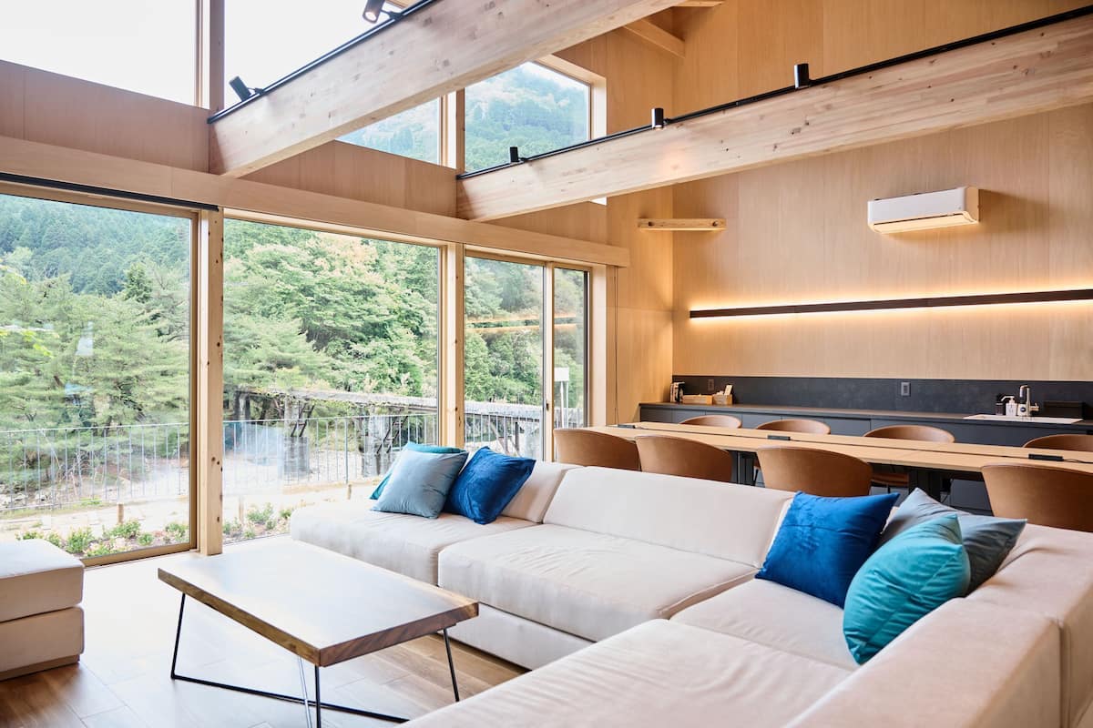 A modern interior with a large sofa, dining table, and chairs, large windows with a view of greenery.