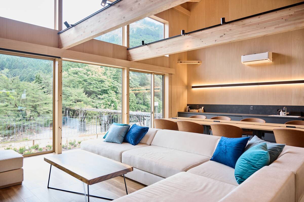 Een modern interieur met een hoekbank, een eethoek en grote ramen met uitzicht op een bebost gebied.
