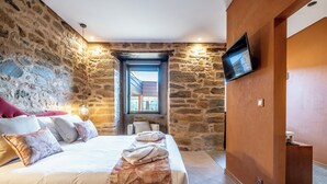 A bedroom with a stone wall, a bed with a floral bedspread, a TV mounted on the wall, and a window with a view. - Cherry Room - Double Superior Room (Paul)
