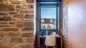 A corner room with a stone wall, a wooden desk, a chair, and a window with a view of buildings. - Cherry Room - Double Superior Room (Paul)