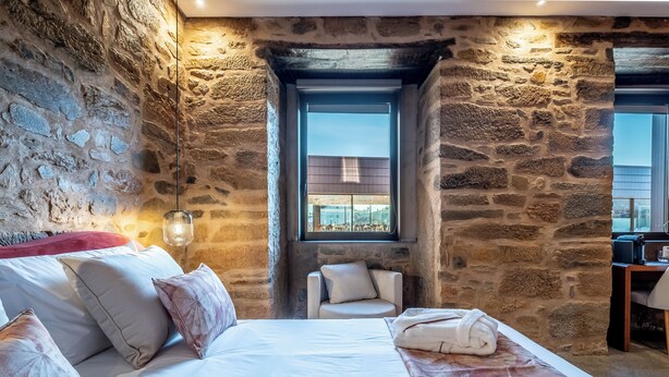A bedroom with stone walls, a bed with white linens, a window with a city view, and a small chair. - Cherry Room - Double Superior Room (Paul)