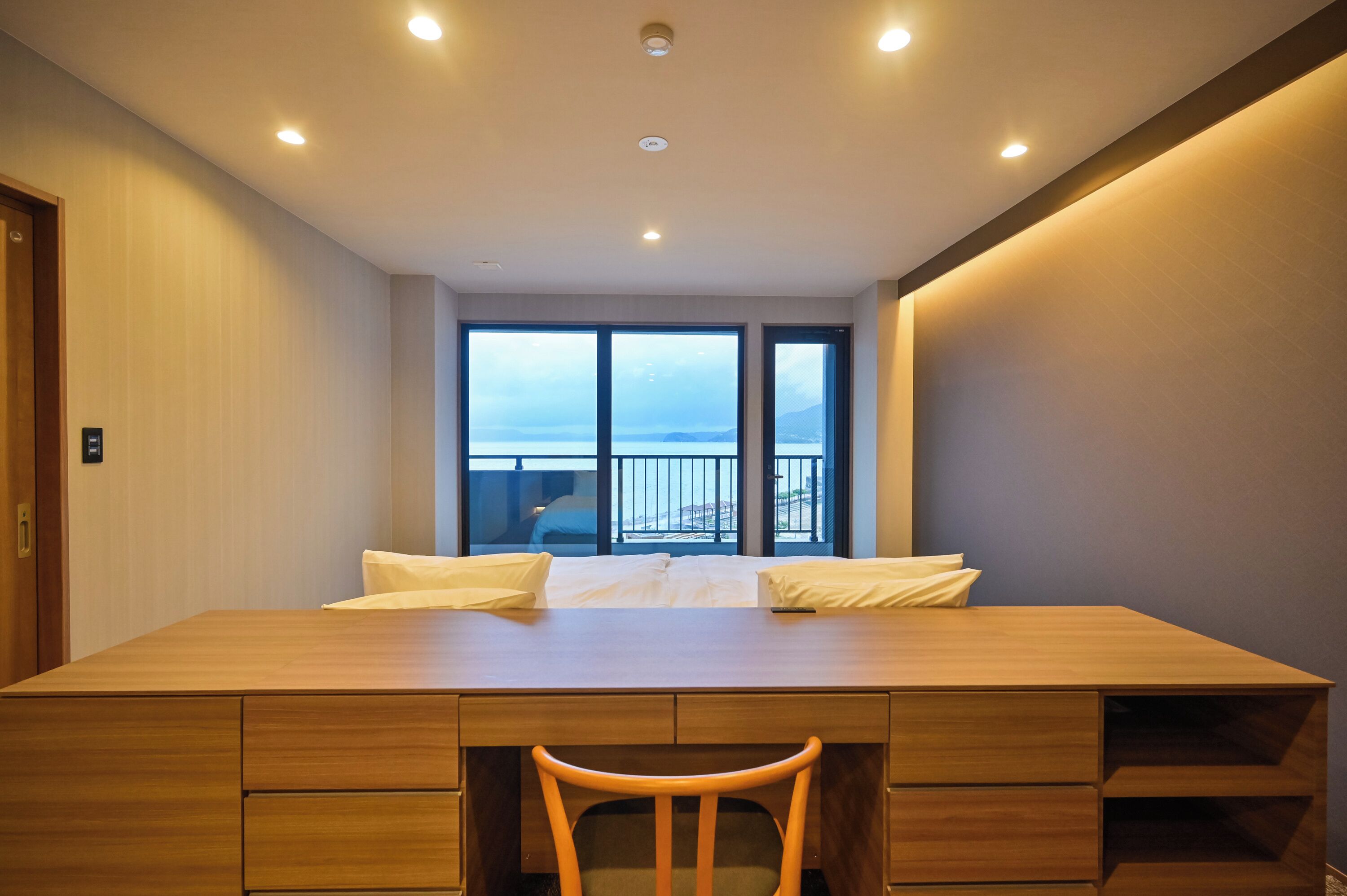Een hotelkamer met een groot houten bureau, twee witte kussens, een houten stoel en uitzicht op zee door grote ramen.