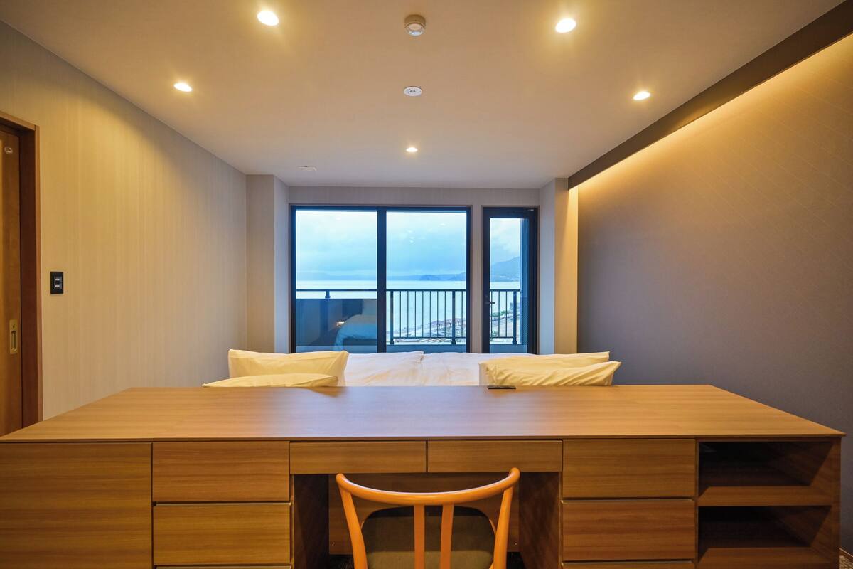 A hotel room with a large wooden desk, two white pillows, a wooden chair, and a view of the sea through large windows.