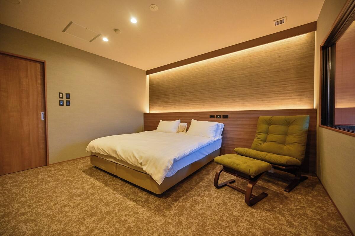 A bedroom with a bed, a green chair, a wooden headboard, and a carpeted floor.