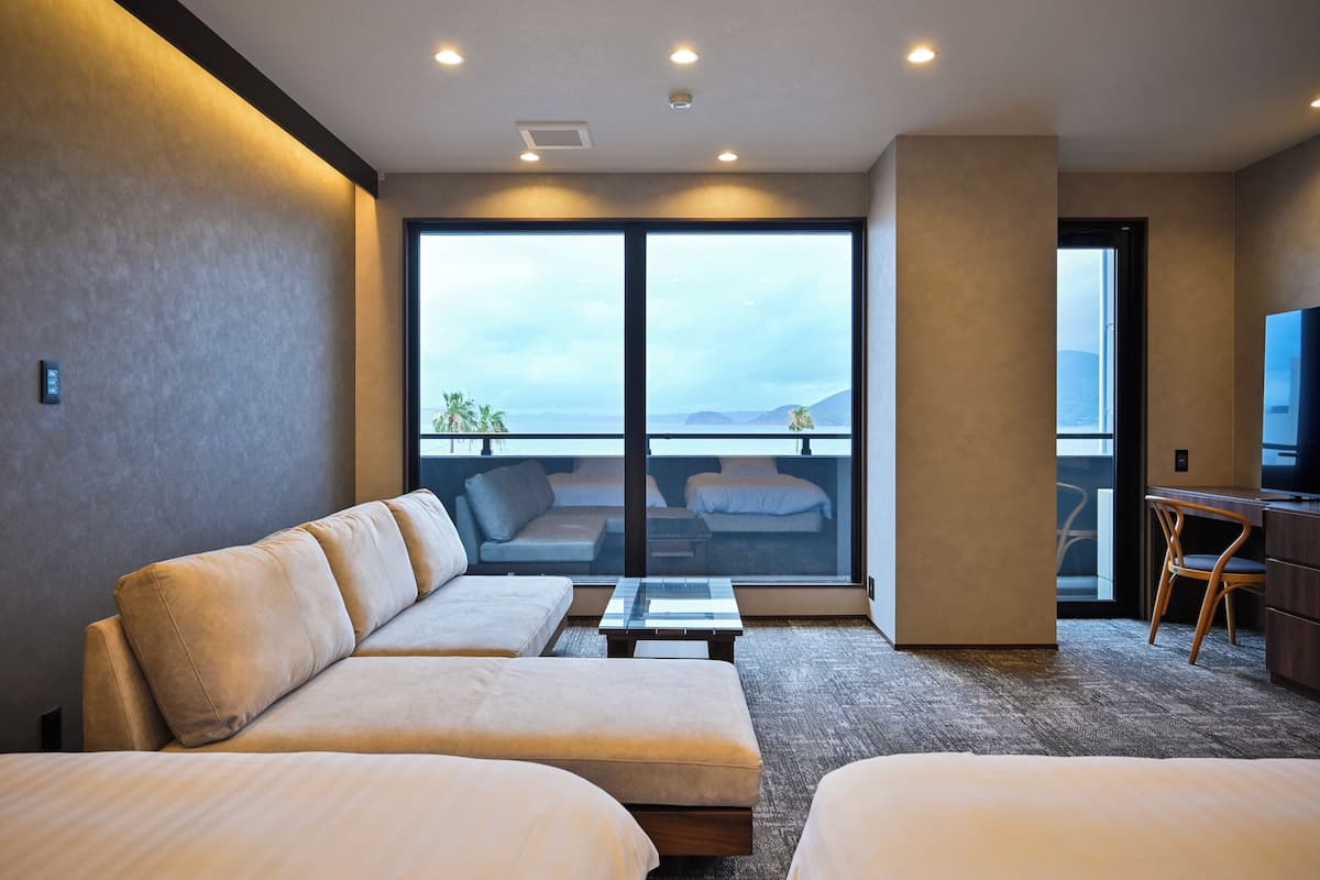 A modern hotel room with a large window, a sofa, a bed, a desk, and a television.