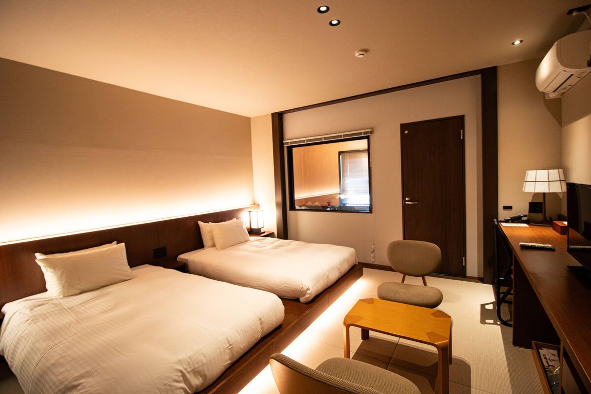 A hotel room with two beds, a desk, a chair, a small table, a mirror, and a door.